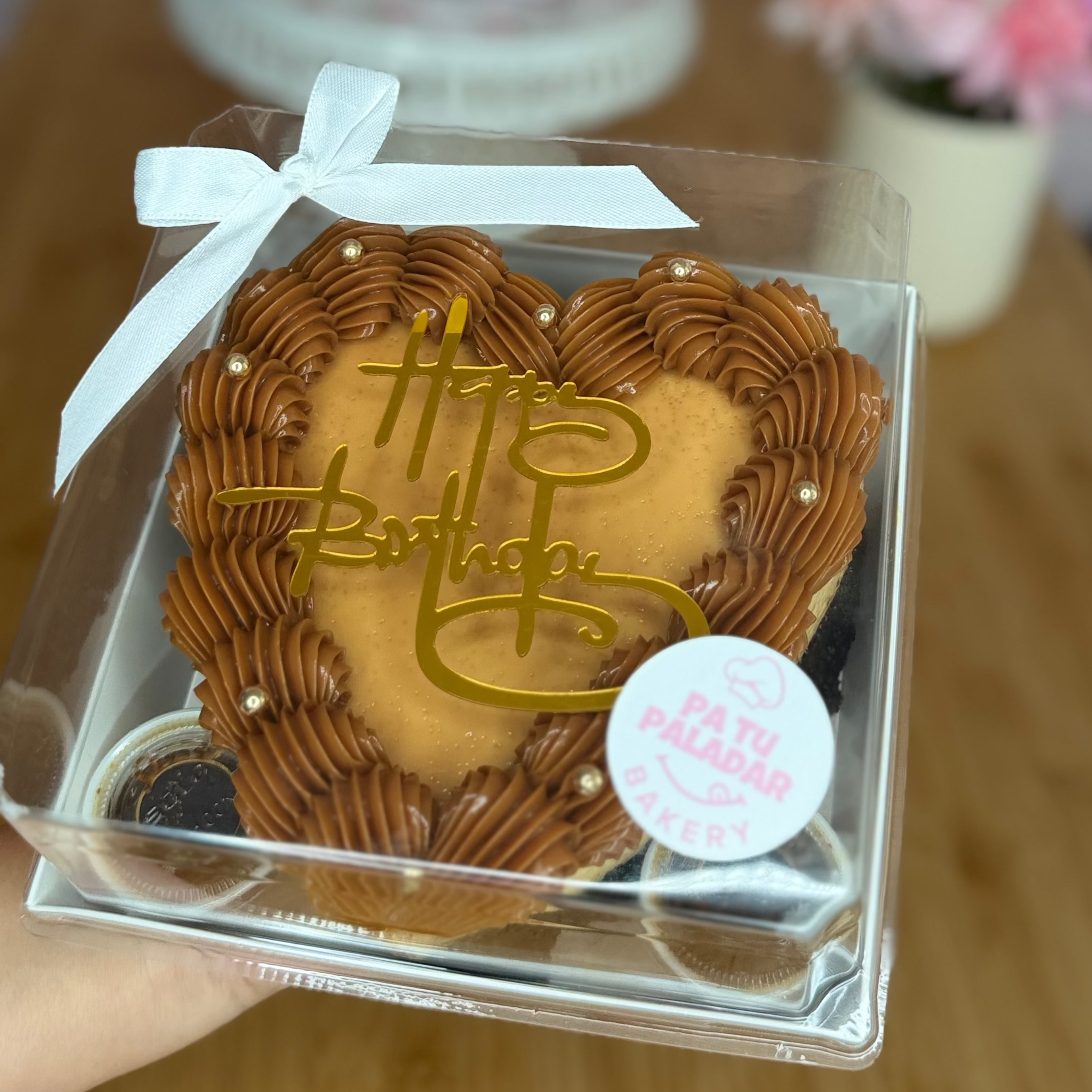 A heart-shaped birthday cake with brown frosting borders, a gold 'Happy Birthday' topper, and a white ribbon bow, packaged in a clear plastic box and held by a hand.